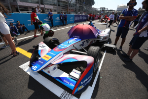 Formula E Rennauto mit weißer, blauer, roter und schwarzer Lackierung auf einer Rennstrecke umgeben von Menschen, mit einem Schirm im Vordergrund und Rennoberfläche im Hintergrund unter einem klaren Himmel.