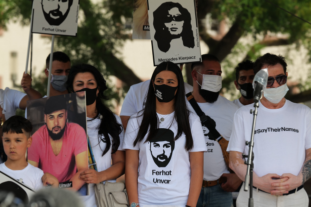 Eine Gruppe von Menschen mit Masken und Schildern bei einer Demonstration in Mexiko-Stadt, mit einem Mikrofon und einer Kamera im Vordergrund und Bäumen und Gebäuden im Hintergrund.