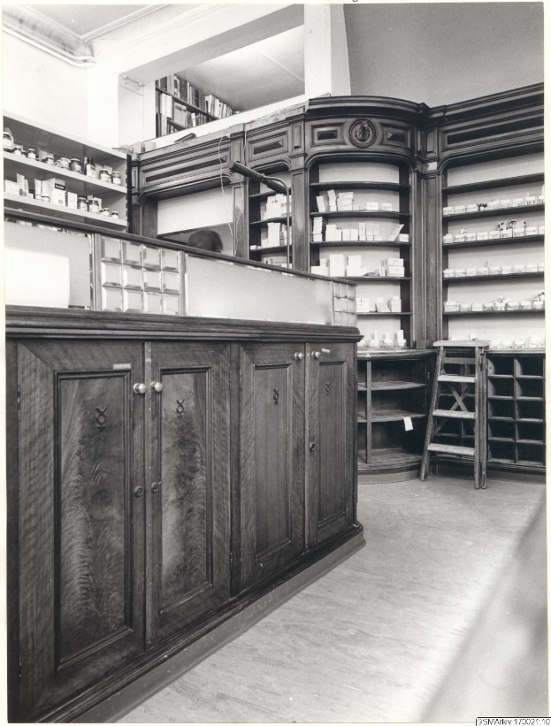 Schwarz-weiß-Foto eines vintage-apothekeninneren mit Theke, Schränken, einer Leiter und Regalen, die mit verschiedenen Gegenständen gefüllt sind, mit Text oben.