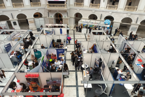 Eine große Gruppe von Menschen versammelte sich in einem Raum voller Stände, Banner, Tische, Stühle und Ballons, mit einem Gebäude im Hintergrund, das Bögen, Säulen, Geländer und einen Bildschirm aufweist.