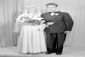 Ein Mann und eine Frau stehen Seite an Seite auf einer Bühne, lächeln und die Frau hält einen Blumenstrauß, mit einem Vorhang im Hintergrund.