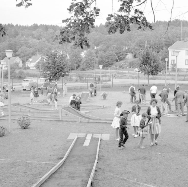 Schwarz-weißes Bild von Menschen, die Minigolf in einem Park mit Grünflächen, Laternen, geparkten Fahrzeugen und Gebäuden im Hintergrund spielen.