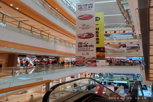 Ein Blick ins Innere eines Einkaufszentrums mit einer Rolltreppe in der Mitte, Menschen, die herumlaufen, einige mit Handtaschen, und Schilder von der Decke hängen, mit Geschäften im Hintergrund.