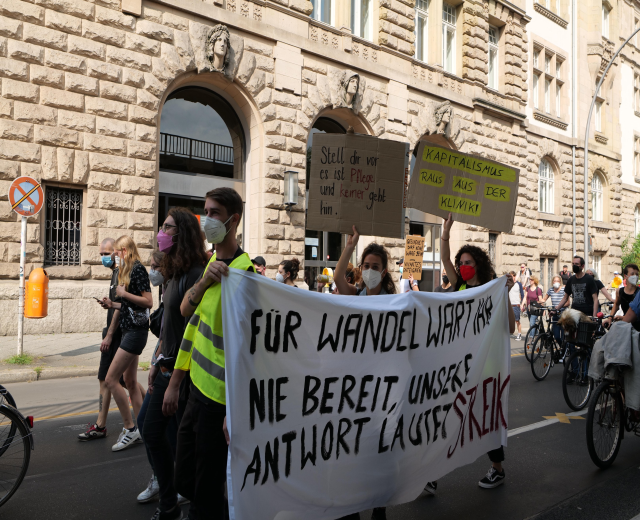 Eine Gruppe von Menschen marschiert bei einer Demonstration auf einer Straße, einige halten Schilder und andere fahren Fahrräder, im Hintergrund ein Gebäude mit Rundbogenfenstern, Säulen und Skulpturen.