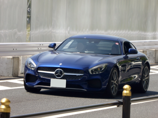 Blauer Mercedes-Benz AMG GT Coupe fährt auf einer Straße mit einer Wand im Hintergrund, mit einem eleganten aerodynamischen Design, eckigen Scheinwerfern, einem großen Kühlergrill, glänzenden großen Rädern und einem luxuriösen Lederinnenraum mit einem modernen Armaturenbrett.