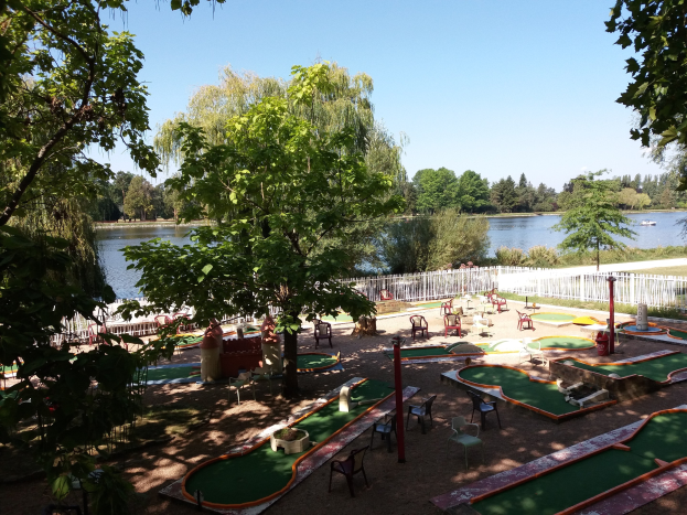 Minigolfanlage in einem Park mit grünem Rasen, Bäumen und einem Zaun, umgeben von verstreuten Stühlen und Tischen, einem Gewässer mit einem Boot im Hintergrund und sichtbarem Himmel.